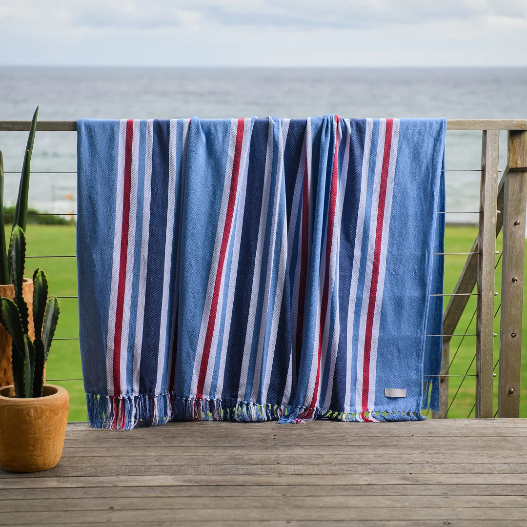 A Protea Pinstripe throw  hanging neatly over a wooden rail. Its hand-knotted fringes and versatile design make it perfect for both indoor and outdoor use. #GuestSatisfaction #UpgradeYourLodge #LodgeLife